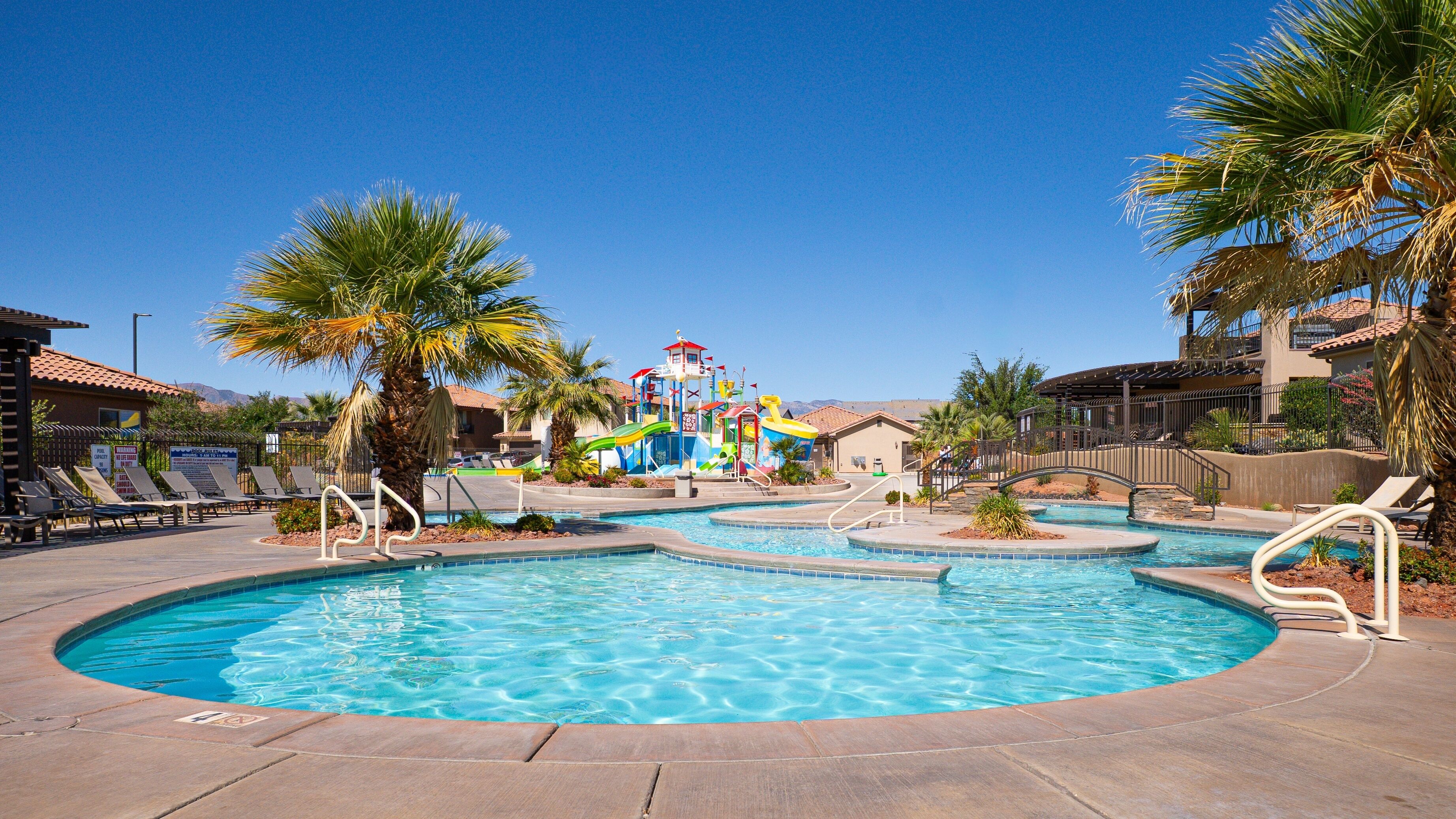 A swimming pool area with multiple pools, lounge chairs, palm trees, and a water park structure in the background.
