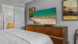 A bedroom with a bed, a wooden dresser, and a large TV mounted on the wall displaying a beach scene.
