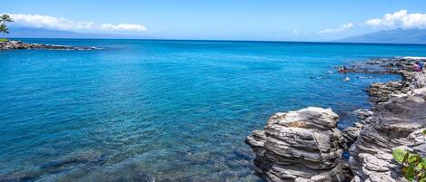 A coastal scene with clear blue water, rocky shore, and a distant mountain.