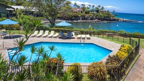 A pool area with lounge chairs and umbrellas, surrounded by a fence and tropical plants, overlooking a coastal view.
