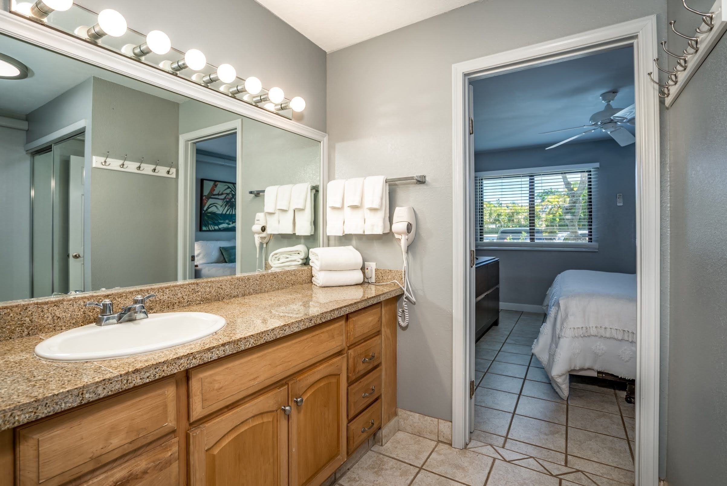 A bathroom with a granite countertop, a white sink, wooden cabinets, a large mirror with lights, a towel rack, and a ceiling fan.