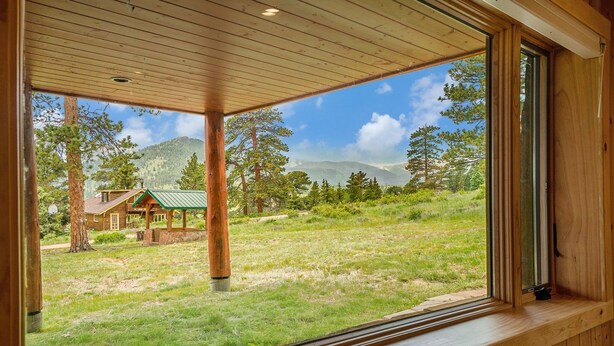 A wooden cabin with a large window overlooking a grassy field and distant mountains.