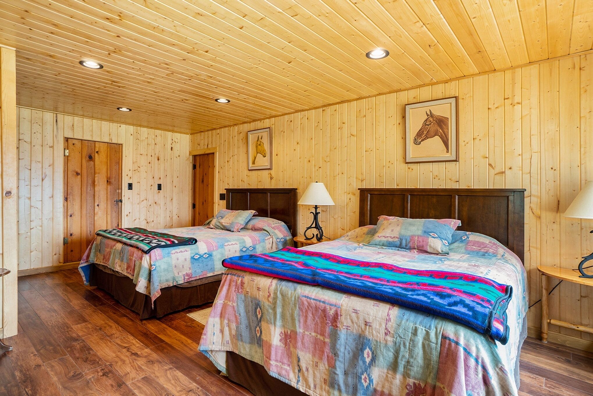 A wooden-paneled room with two beds, each with a colorful blanket, a nightstand with a lamp, and a framed horse picture on the wall.