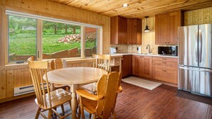 A wooden-paneled kitchen with a dining table and chairs, a refrigerator, and a view of a grassy yard through large windows.