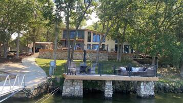 Une propriété au bord du lac, dotée d’une jetée en pierre, d’un espace salon extérieur et d’une grande maison à plusieurs niveaux et avec des balcons.