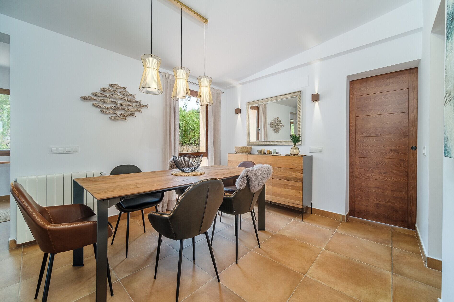 A dining area with a wooden table, four chairs, pendant lights, a mirror, and a wooden door.