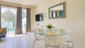 Un coin repas avec une table en verre, des chaises blanches, une plante en pot et un téléviseur à écran plat fixé au mur.