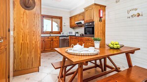 A kitchen with wooden cabinets, a dining table with a tray of white dishes, and a bowl of green apples. - Binifortu - Four Bedroom Villa, Sleeps 8 (Punta Prima)