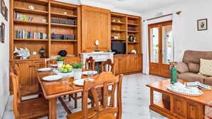 A spacious living room with a wooden dining table, chairs, and a sofa. There are bookshelves filled with books and decorative items. A television is mounted on the wall, and a door leads to an outdoor area. - Binifortu - Four Bedroom Villa, Sleeps 8 (Punta Prima)