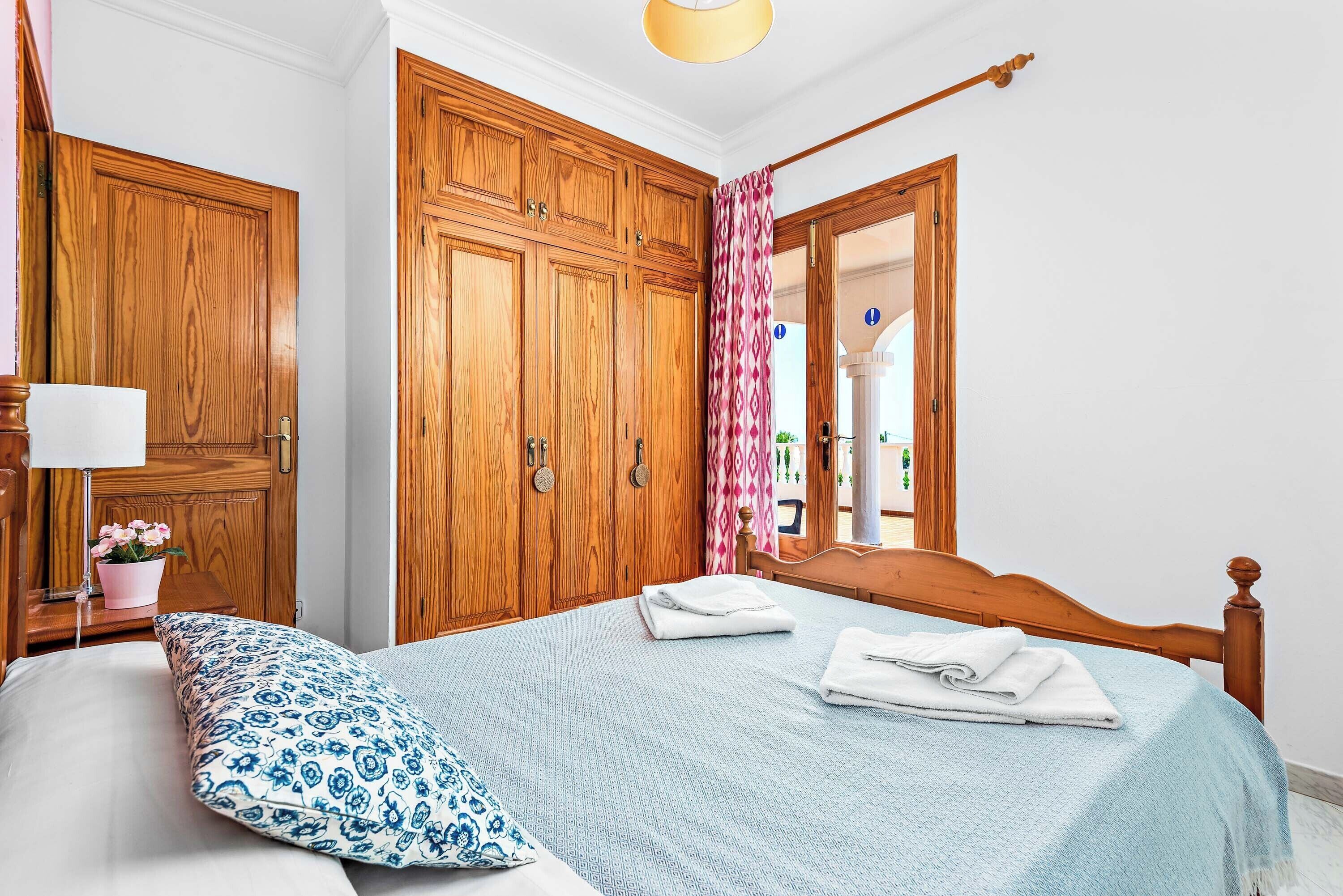 A bedroom with a bed, wooden wardrobe, and a nightstand with a lamp and flowers.