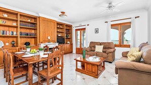 A spacious living room with a wooden dining table, a sofa, and a TV. There are bookshelves and a ceiling fan. - Binifortu - Four Bedroom Villa, Sleeps 8 (Punta Prima)