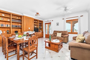 A spacious living room with a wooden dining table, a sofa, and a TV. There are bookshelves and a ceiling fan.