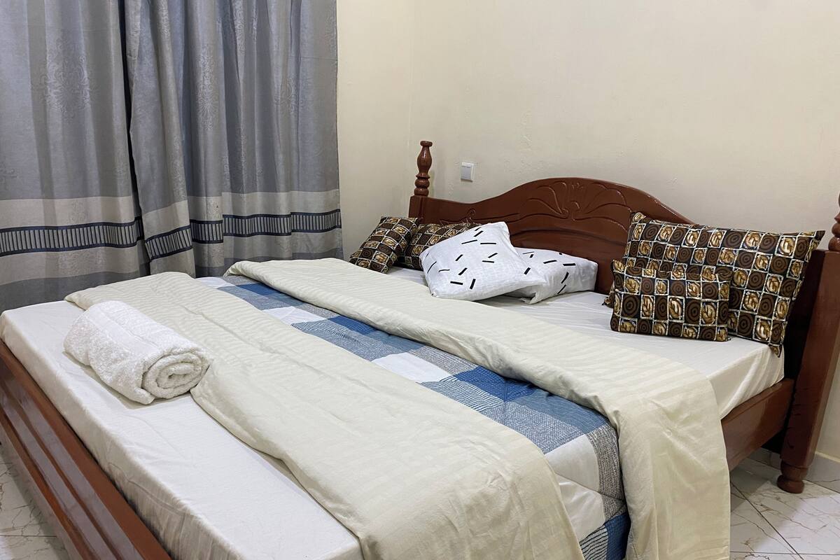A neatly made bed with a white and blue quilt, a rolled white towel, and patterned pillows.