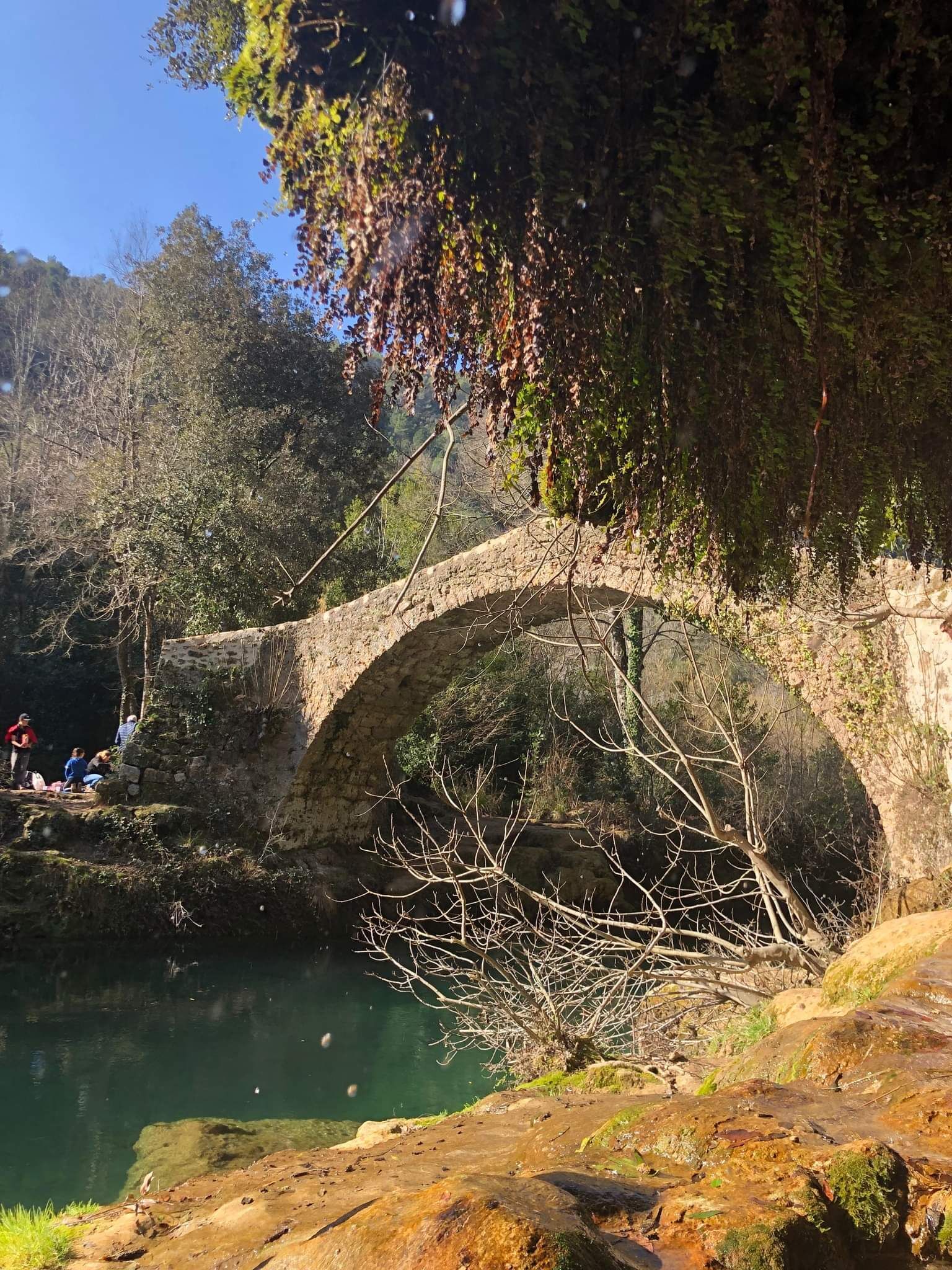Un pont de pierre enjambe une rivière, et des personnes sont assises sur la rive.