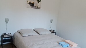 A neatly made bed with a beige comforter and pillows, a bedside table with a tissue box, a wall-mounted lamp, and a hanging light fixture.