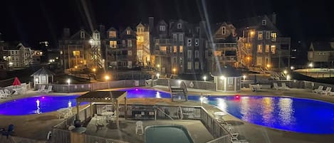 A nighttime view of a resort with two large swimming pools, a covered deck area, and a series of multi-story buildings with balconies.