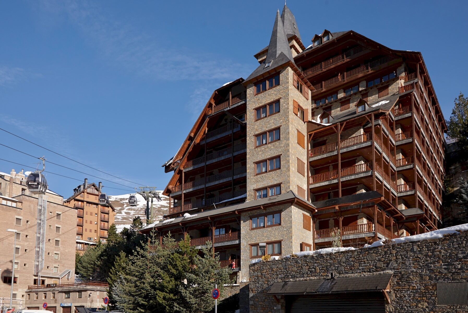 A multi-story hotel with a stone facade, wooden balconies, and a ski lift in the background.