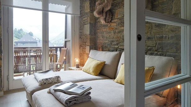 A bedroom with a stone wall, a large window, a bed with pillows, a bedside table with a lamp, and a view of a balcony with chairs. - Port Luxe by FeelFree Rentals (Baqueira)