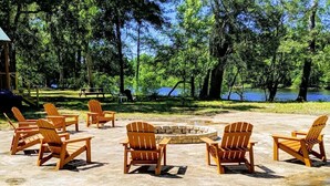 Uma fogueira rodeada por cadeiras de madeira Adirondack em um ambiente natural, com árvores e um corpo d'água ao fundo.