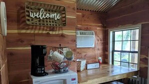 Interior de uma cabana de madeira com um letreiro de boas-vindas, um micro-ondas branco, uma cafeteira e uma janela com vista.