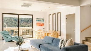 A modern living room with a sofa, a dining area with chairs, and a view of a forested hillside through large windows.