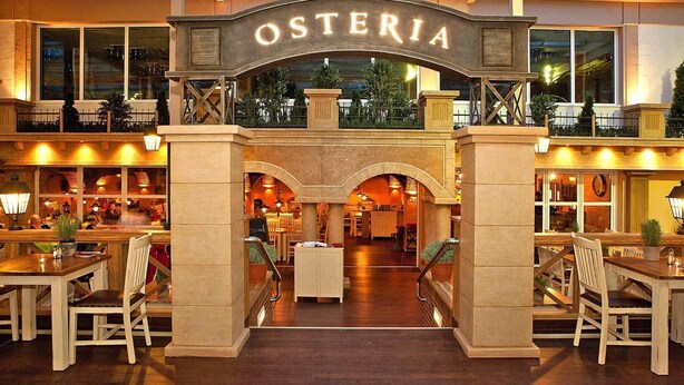 Een hotel lobby met een centrale boog waarop 'OSTERIA' staat, met een bar aan de linkerkant en een zithoek aan de rechterkant.