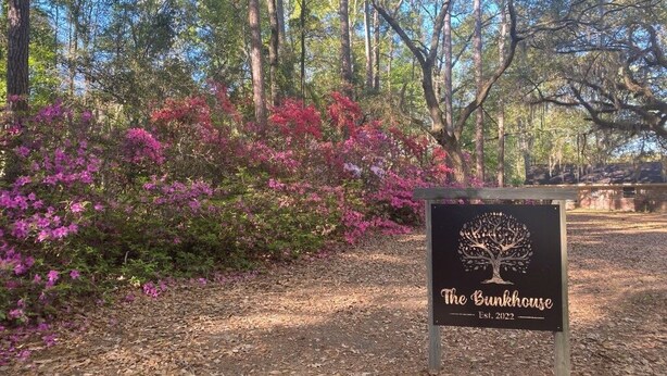 Schild mit dem Schriftzug 'The Bungalow' und einem Baum-Logo, umgeben von blühenden rosafarbenen Sträuchern.