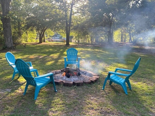 Un hogar para fuego con cuatro sillas Adirondack azules dispuestas alrededor en un área verde con árboles al fondo.