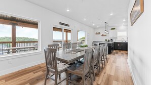A spacious dining area with a wooden table and chairs, large windows offering a view of the outdoors, and a kitchen area with modern appliances.