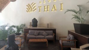 A room with a sofa, a wooden table, and a Buddha statue. The wall has the words "ALDEA THAI" illuminated.