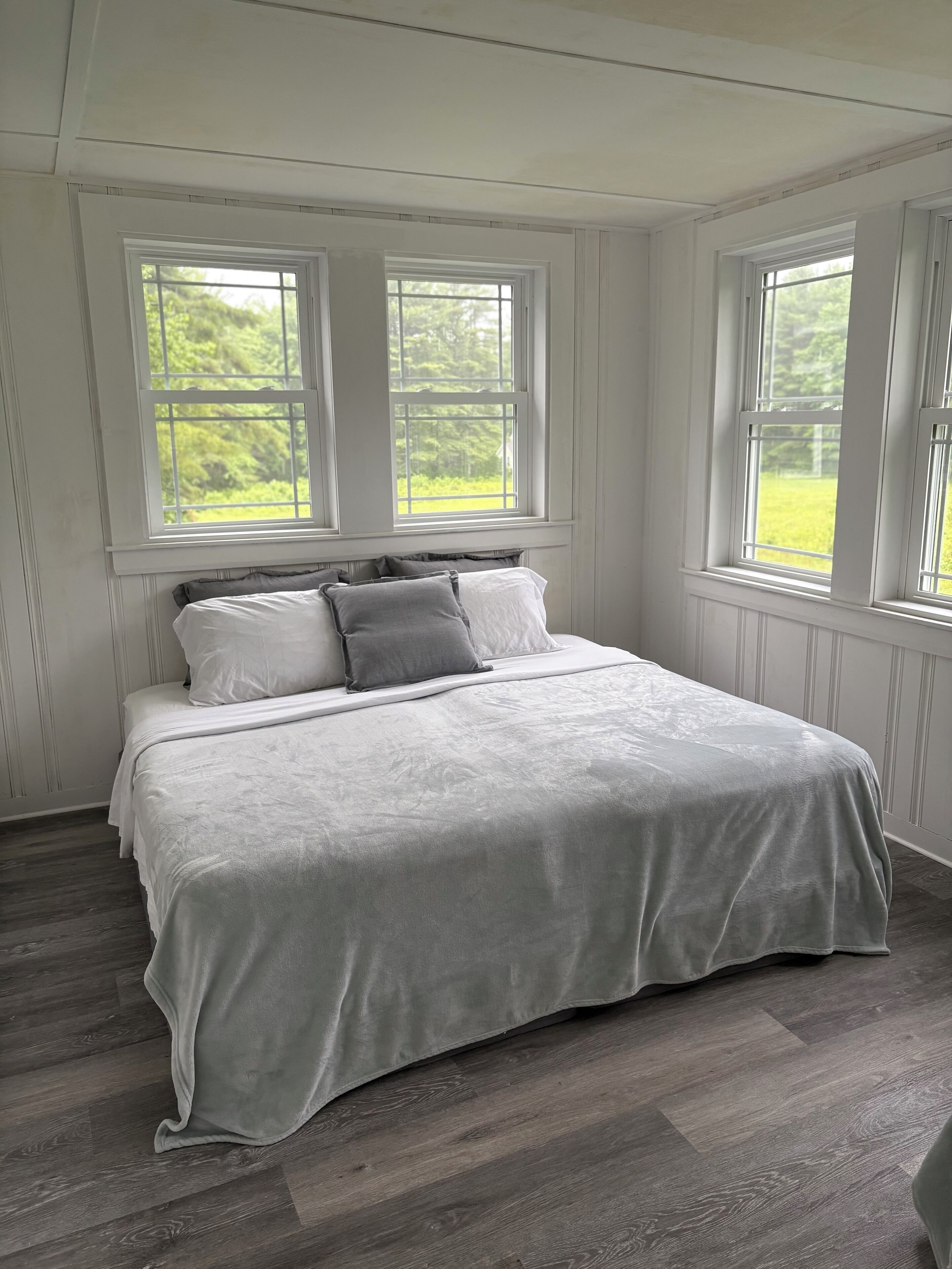 A bedroom with a large bed, two windows, and a view of greenery outside.