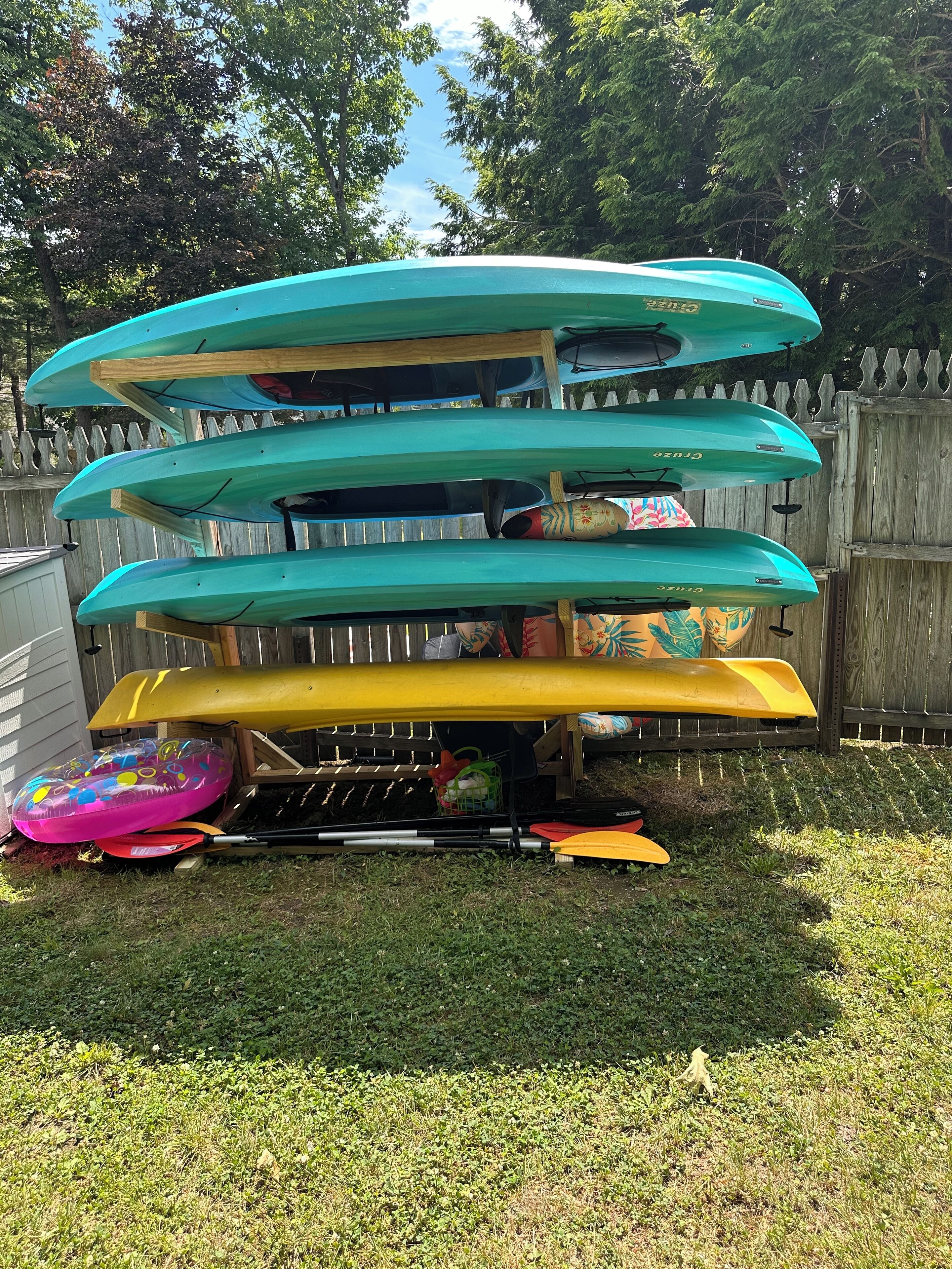 Une pile de kayaks et une planche de paddle sont posées dans un jardin, avec une clôture en bois et de l’herbe.