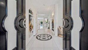 Un pasillo con puertas negras, paredes blancas y piso de baldosas que conduce a una habitación con cama y una silla.