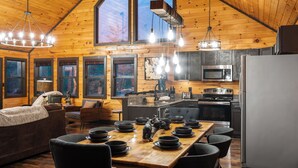Un interior de cabaña acogedor con una mesa de comedor de madera dispuesta para cuatro personas, una cocina con electrodomésticos modernos y un gran ventanal que proporciona luz natural.