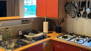 A compact kitchen with a gas stove, a sink, and a window with curtains. - Mobil Home 4/6 Clim Terrasse Barbecue Piscines Toboggan Animations (Le Muy)