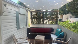 A covered patio with a wooden deck, a red cushioned sofa, and two white cushioned chairs. - Mobil Home 4/6 Clim Terrasse Barbecue Piscines Toboggan Animations (Le Muy)