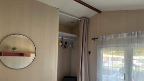 A room with a wooden wardrobe, a mirror, a window with sheer curtains, and a shelf with a small plant. - Mobil Home 4/6 Clim Terrasse Barbecue Piscines Toboggan Animations (Le Muy)