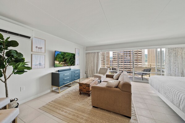 A modern hotel room with a large bed, a sofa, a TV, a desk, and a balcony with a city view.