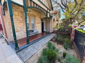 A building with a stone facade, a covered porch with a bench, and a small garden with various plants.