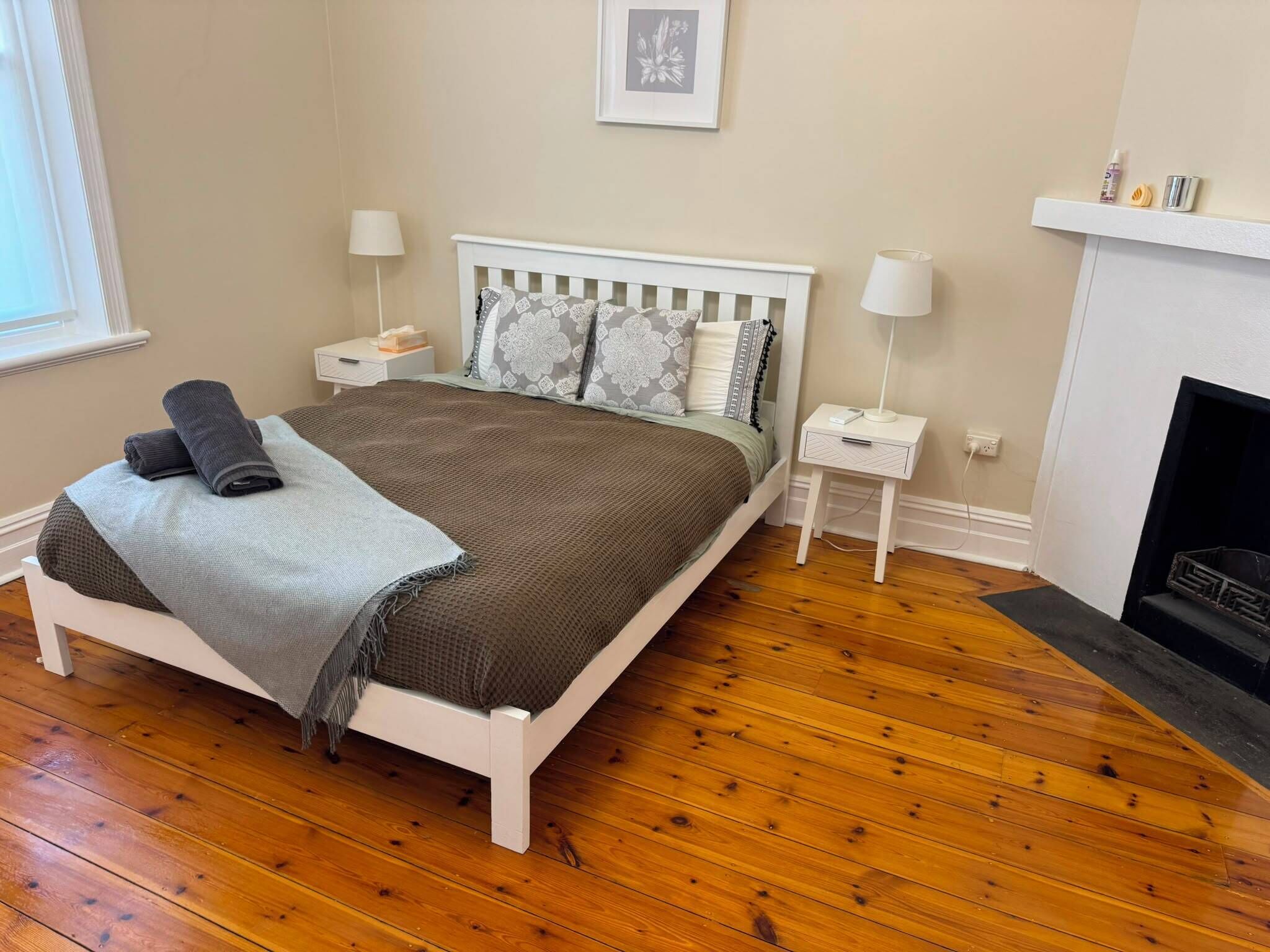 A bedroom with a white bed, wooden flooring, a bedside table with a lamp, and a fireplace.