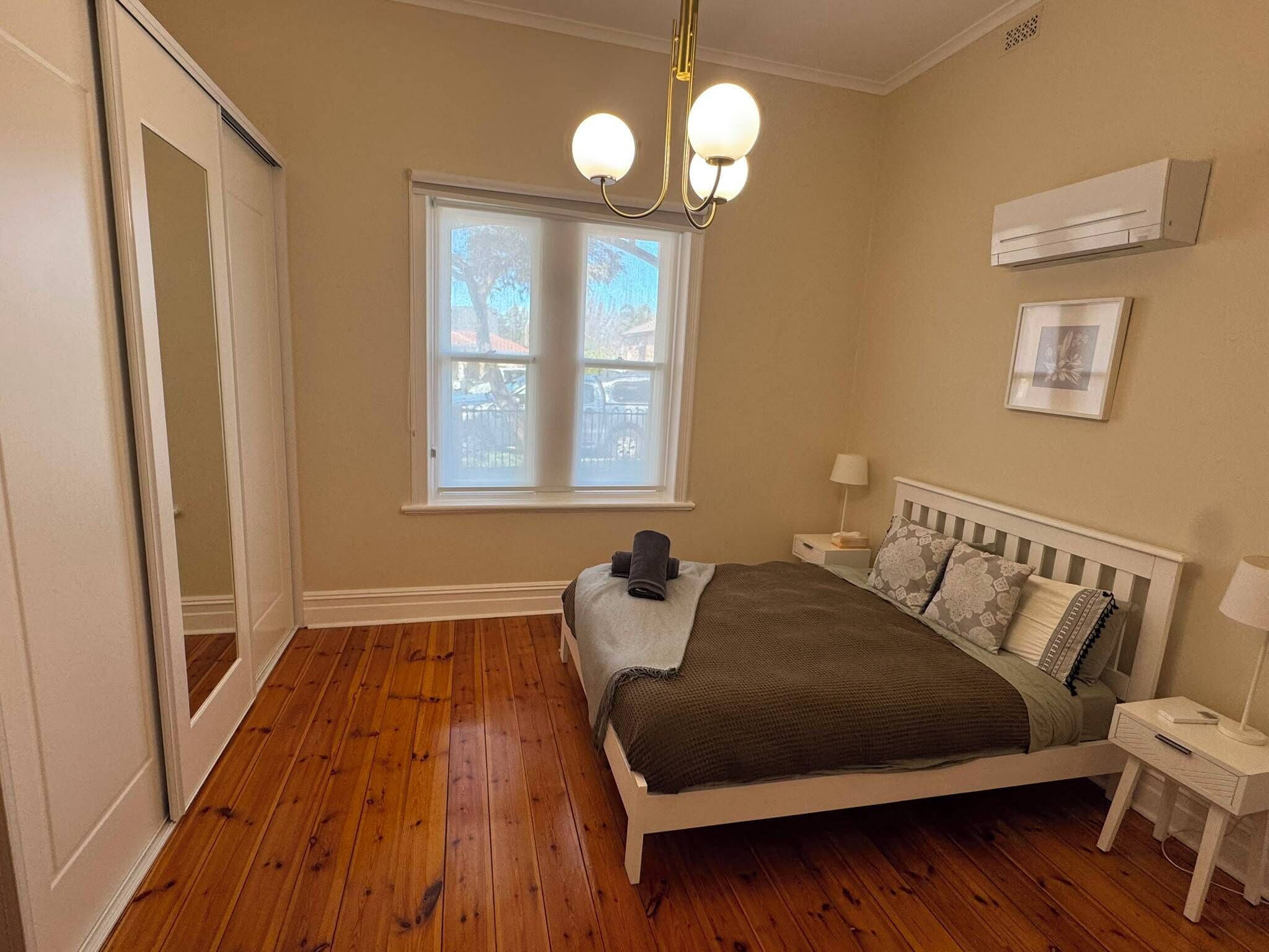 A bedroom with a wooden floor, a bed with a white headboard, a window with blinds, a ceiling light fixture, and an air conditioning unit.
