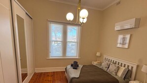 A bedroom with a wooden floor, a bed with a white headboard, a window with blinds, a ceiling light fixture, and an air conditioning unit.