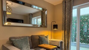 A modern living room with a sofa, a small table, a large mirror, and a view of a garden through a sliding glass door. - Central Studios (Cardiff)