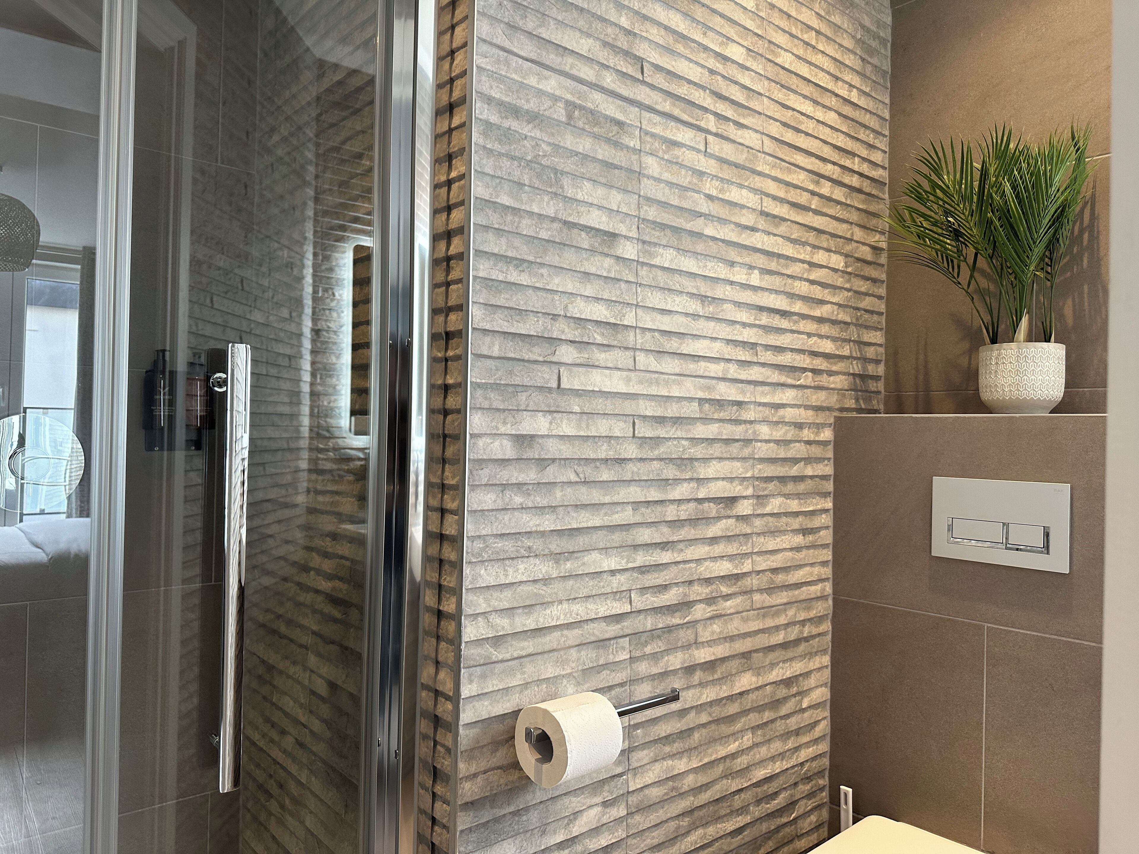 Een moderne badkamer met een douchecabine van glas, een structuurwand, een toiletrolhouder en een plant in pot.