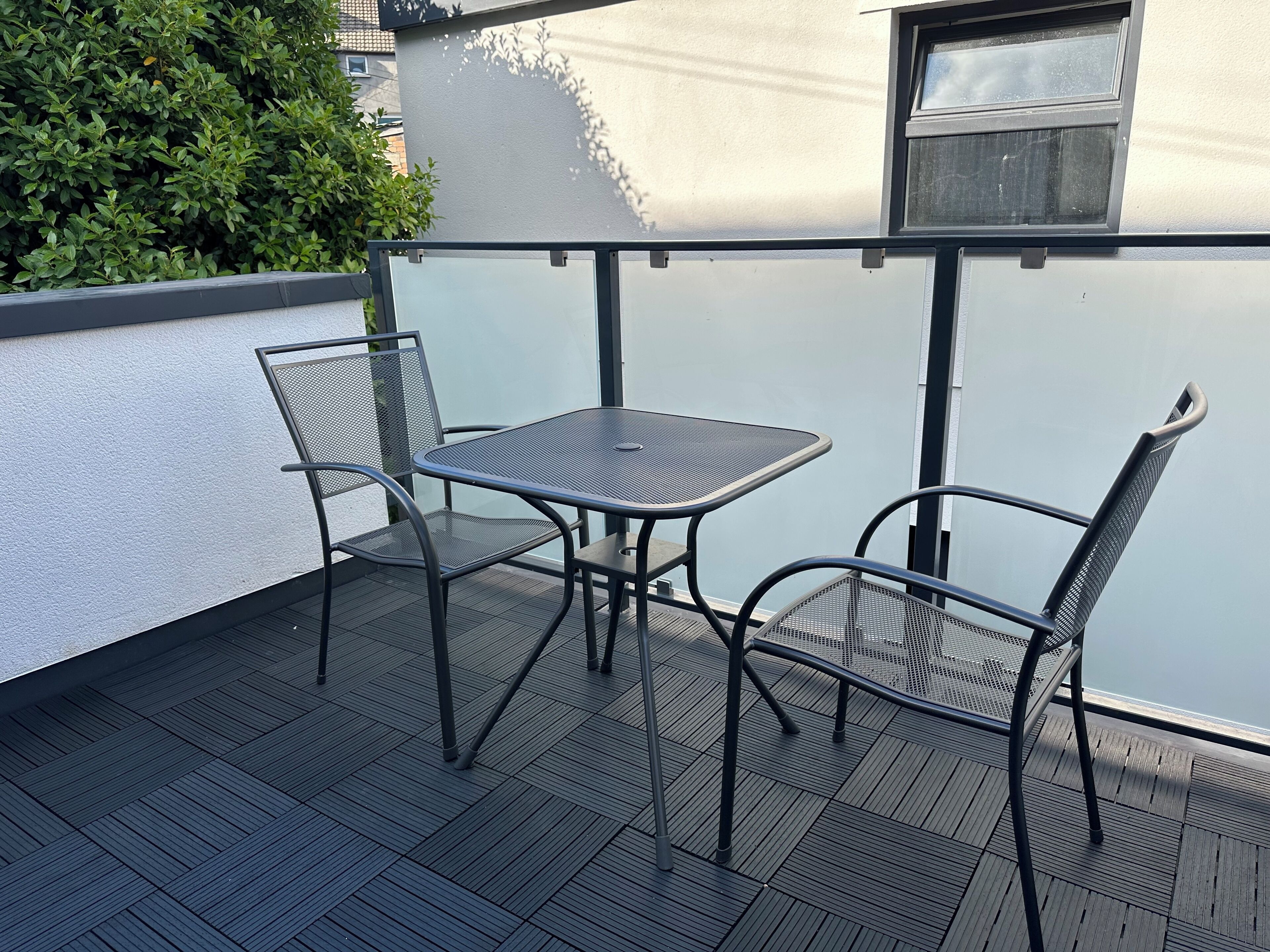 A balcony with a metal table and chairs, a window, and a white wall.