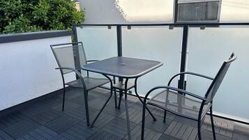 A balcony with a metal table and chairs, a window, and a white wall.