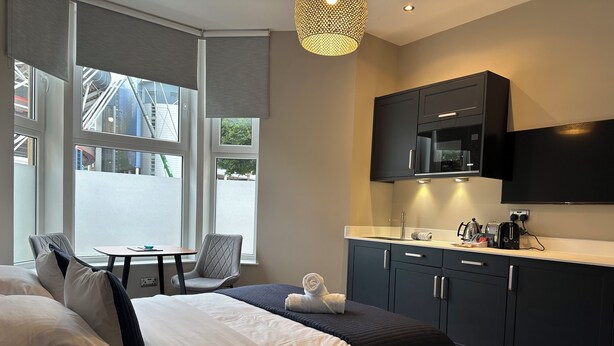 A modern hotel room with a bed, a small table, a chair, a kitchenette, and large windows. - Central Studios (Cardiff)