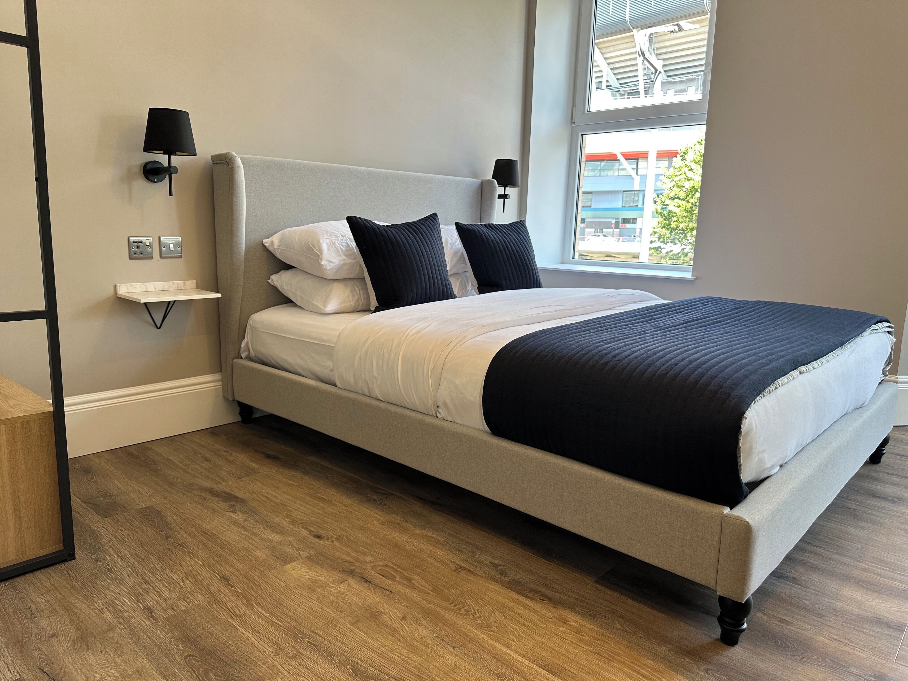 A neatly made bed with a headboard, two bedside lamps, a small shelf, and a window with a view of buildings and trees.