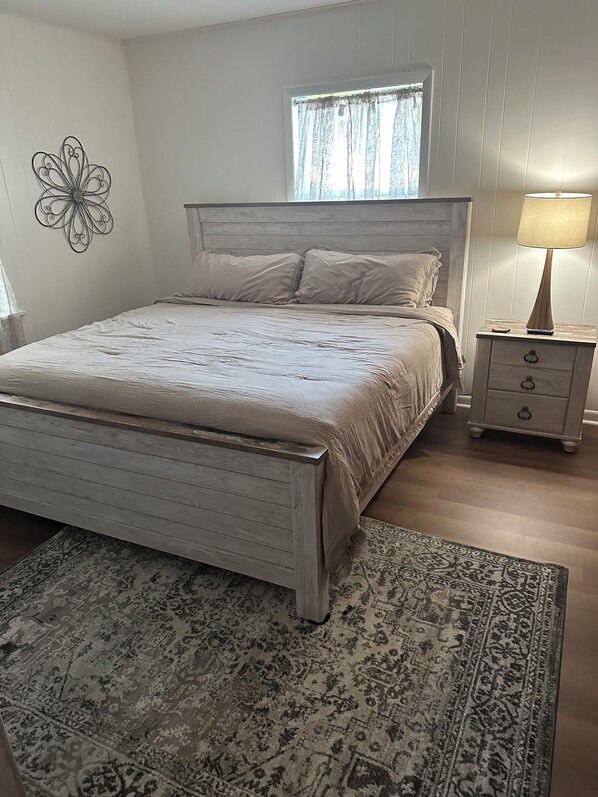 Un dormitorio con cama, mesita de noche, lámpara, ventana con cortinas y un adorno en la pared.