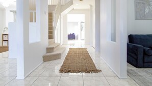 A spacious hallway with a staircase leading to an upper level, a blue sofa, and a large woven rug on the floor. - Spacious, Sunlit & Recently Renovated Family Home - 4BD & Hotel Quality Bedding (Prestons)